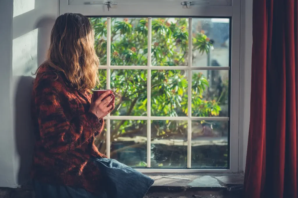 Woman looking out window with a mug, supporting mental health and anxiety treatment.